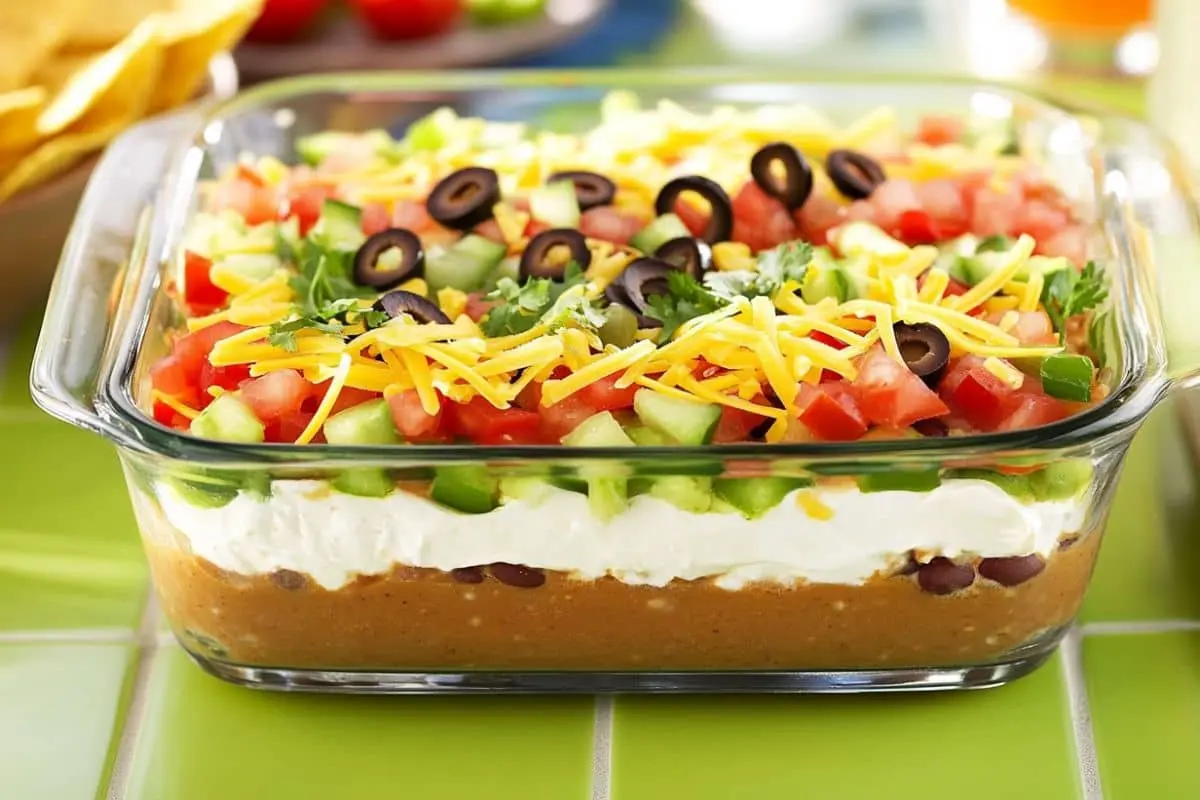 A glass dish filled with seven-layer taco dip, featuring layers of refried beans, sour cream, diced vegetables, shredded cheese, and sliced black olives.