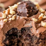 A close-up of a chocolate brownie cupcake with creamy frosting, a bite taken out, and chocolate shavings on top.