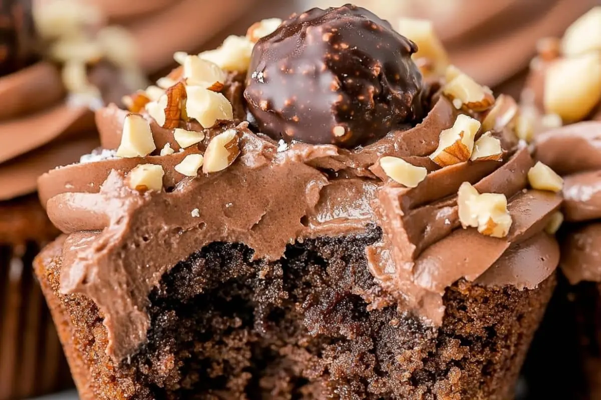 A close-up of a chocolate brownie cupcake with creamy frosting, a bite taken out, and chocolate shavings on top.