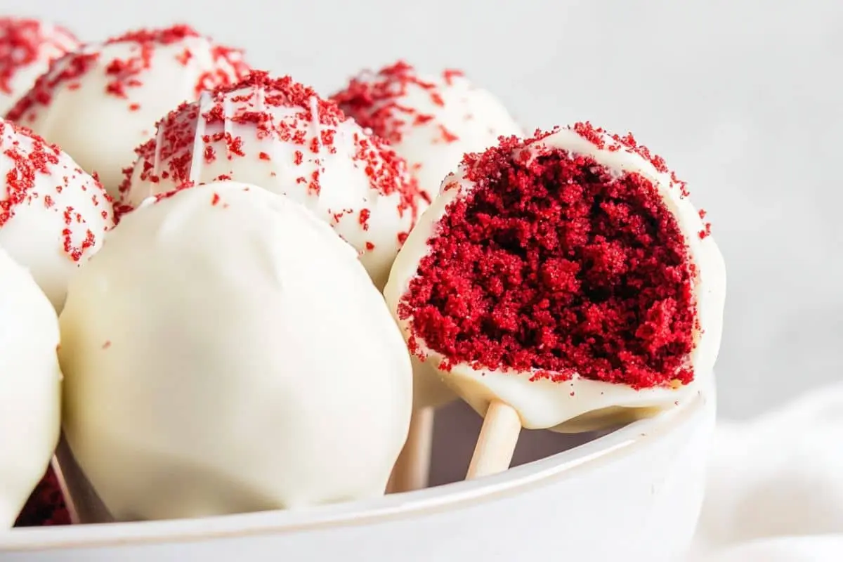 A close-up of red velvet cake pops coated in white chocolate, with one cut open to reveal the moist red cake inside.