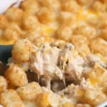 A spoonful of creamy chicken tater tot casserole with melted cheese being lifted from a baking dish.